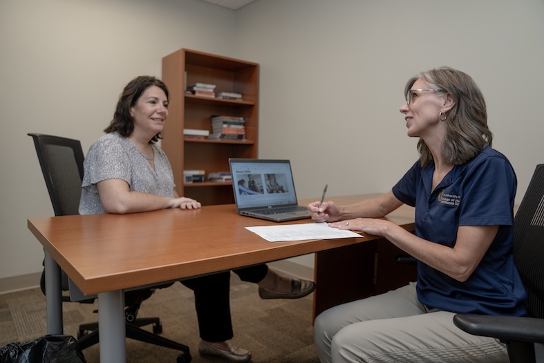 Two women sit across from each other at a desk in an office. One woman is taking notes on a clipboard, while the other is smiling. A laptop and bookshelf are visible in the background.
